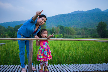 Happy Mother and her child play outdoors having fun, and pointing at something in the Green rice field.