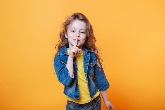 Cute curly blonde girl looking camera and making silence gesture isolated over yellow background