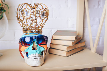 A human skull in sunglasses with a crown and books on the shelf.