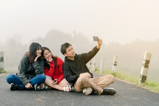 group of three Asian friends wearing a sweater using a selfie phone to take pictures in a tourist attraction. Along the road beside the reservoir in the thick fog With a smiling cof happiness