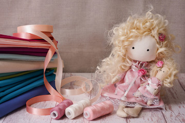 Cute handmade doll on a wooden table near colorful fabrics, knitted lace, pastel ribbons
