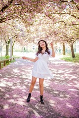 A young beautiful Asian woman in a white dress walks in a flowered park. Sakura. Blooming trees.
