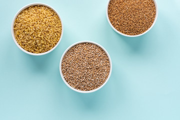 Uncooked crushed spelt, wheat and bulgur in a white bowl on pastel blue background. Top view, copy space, minimal idea