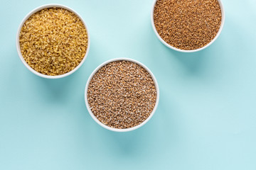 Uncooked crushed spelt, wheat and bulgur in a white bowl on pastel blue background. Top view, copy space, minimal idea