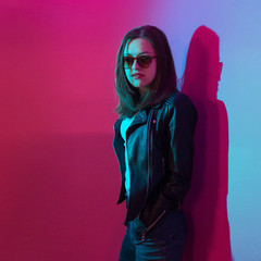 Fashioned girl in leather jacket in dark glasses in pink and blue  neon lights background. 