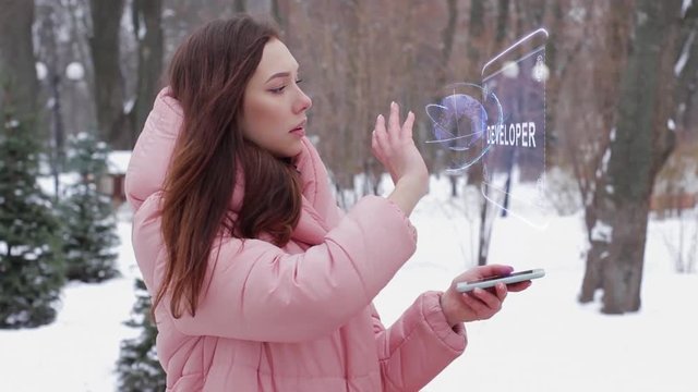 Beautiful young woman in a winter park interacts with HUD hologram with text Developer. Red-haired girl in warm pink clothes uses the technology of the future mobile screen