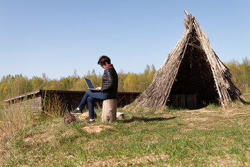 Girl using lap top among nature. Time online. Is sitting on a stump near a grass hut on the Sunset.