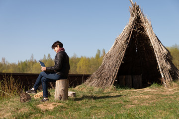 Girl using lap top among nature. Time online. Is sitting on a stump near a grass hut on the Sunset.