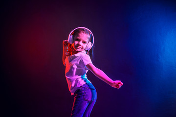 Fototapeta na wymiar Girl of 7 years old listening to music in headphones and jump on dark colorful background. Neon light. Dancing girl. Happy small girl dancing to music. Cute child enjoying happy dance music.