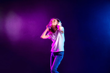 Girl listening to music in headphones on dark colorful background. Cute child enjoying happy dance music, close eye's and smile posing on studio background wall.