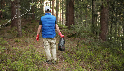 Adult man volunteer in red gloves removes plastic trash in forest, caring for environment.