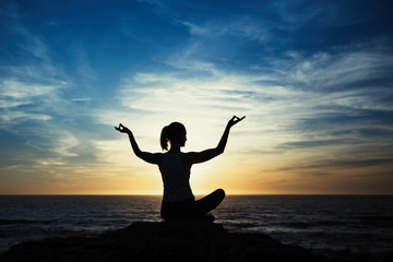 Yoga woman silhouette sitting in Lotus position on the shore of the evening ocean.