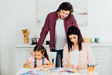 handsome father standing and looking at brunette wife and daughter drawing at home