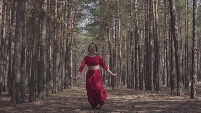 Skilled graceful woman in red dress dancing in the forest landscape. Beautiful contemporary dancer. Graceful girl runs and jumps. The camera moves in parallel with the girl. Shooting from the side.
