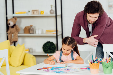 handsome father standing near cute daughter drawing at home