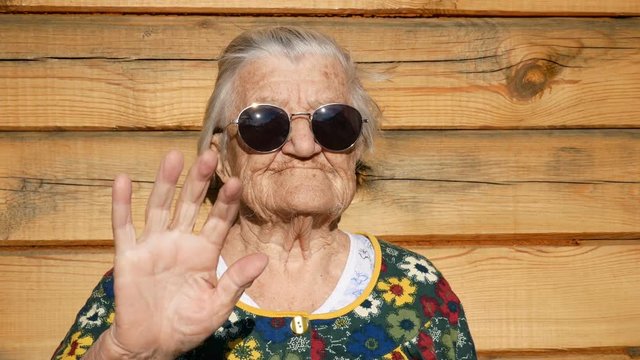 Brutal granny in sunglasses waves by hand at the camera.  Wrinkled face of aged woman. Grandmother on wooden rustic background. Old granny looks into the camera and waves by hand. 91 year old grandma.