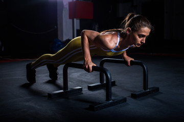 Fototapeta na wymiar Young athletic girl in yellow tight sports uniform doing horizontal push-ups with bars in gym