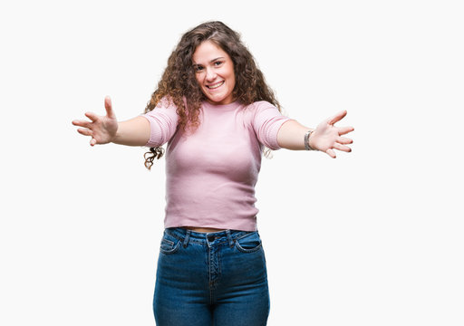 Beautiful brunette curly hair young girl wearing pink sweater over isolated background looking at the camera smiling with open arms for hug. Cheerful expression embracing happiness.