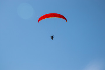 Paramotor (powered paraglider) with red parachute flying in blue sky. Back view with some flare.