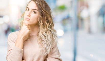 Beautiful young blonde woman wearing sweatershirt over isolated background with hand on chin thinking about question, pensive expression. Smiling with thoughtful face. Doubt concept.