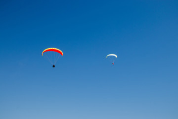 Two paramotors (powered paragliders) with red and yellow parachutes flying in blue sky.