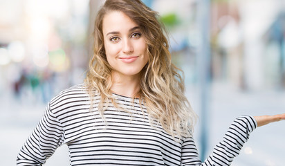 Beautiful young blonde woman wearing stripes sweater over isolated background smiling cheerful presenting and pointing with palm of hand looking at the camera.