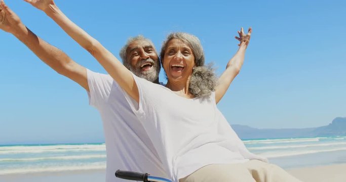 Front view of active senior African American woman sitting on man bicycle handlebars at beach 4k