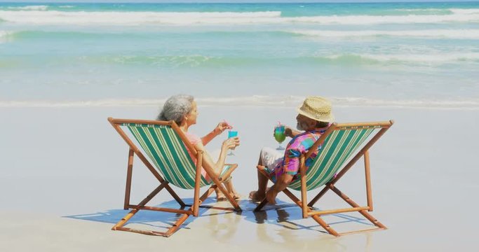 Rear view of active senior African American couple toasting drinks on deckchair at beach 4k
