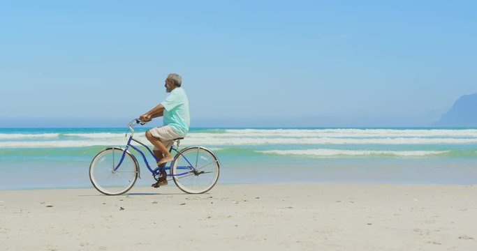Side view of active senior African American man riding bicycle on beach in the sunshine 4k