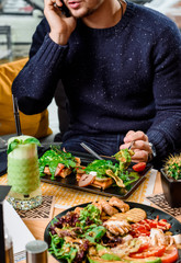 Man eats delicious savory breakfast - waffles with ham, cheese, eggs, tomatoes and spinach sauce and drinks a green smoothie in a restaurant. Waffles on a black plate on a wooden table in restaurant.