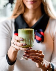 Young girl in a light sweater holds a beautiful faceted glass of green matcha latte, sprinkled with nut chips. close-up. Concept - healthy food