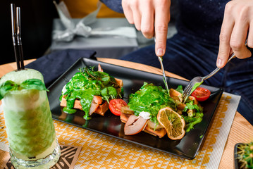 Man eats delicious savory breakfast - waffles with ham, cheese, eggs, tomatoes and spinach sauce and drinks a green smoothie in a restaurant. Waffles on a black plate on a wooden table in restaurant.