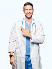 Handsome doctor man wearing medical uniform over isolated background cheerful with a smile of face pointing with hand and finger up to the side with happy and natural expression on face