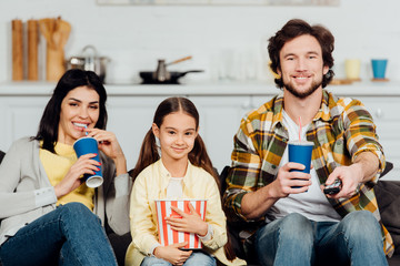 happy parents holding disposable cups near cheerful daughter with bucket of popcorn