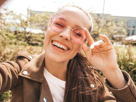 Closeup portrait of beautiful smiling brunette girl in summer hipster jacket. Model taking selfie on smartphone.Woman making photos in warm sunny day in the street in sunglasses