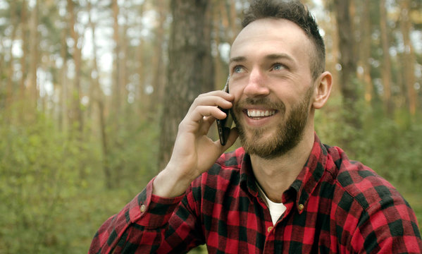 Handsome young bearded man has pleasant talk via smartphone while relaxing in the pine forest