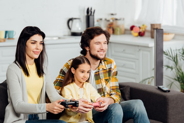 happy man sitting near cheerful kid and wife and playing video game at home