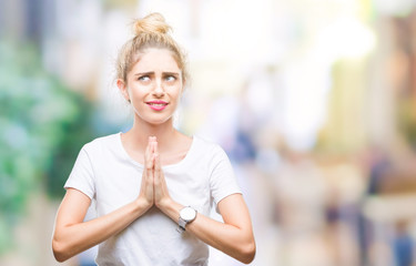 Young beautiful blonde woman wearing white t-shirt over isolated background begging and praying with hands together with hope expression on face very emotional and worried. Asking for forgiveness