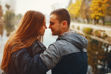 Cute couple sitting in a autumn park. Boy in a black jacket. Girl in a gray dress. Couple sitting near river