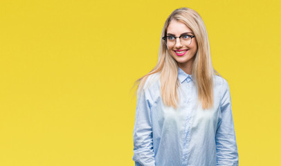 Young beautiful blonde business woman wearing glasses over isolated background looking away to side with smile on face, natural expression. Laughing confident.