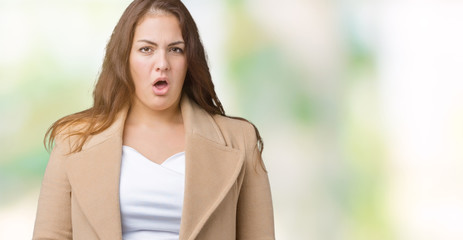 Beautiful plus size young woman wearing winter coat over isolated background In shock face, looking...