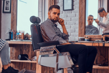 Attractive pensive man is waiting for his turn to get a haircut at busy barbershop.