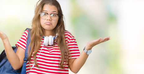 Fototapeta na wymiar Young beautiful brunette student woman wearing headphones and backpack over isolated background clueless and confused expression with arms and hands raised. Doubt concept.