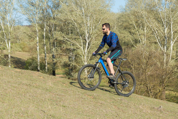 Naklejka premium Cyclist in shorts and jersey on a modern carbon hardtail bike with an air suspension fork rides off-road on green hills near the forest 