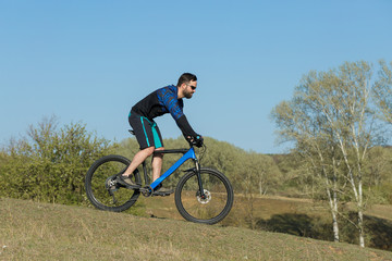 Fototapeta na wymiar Cyclist in shorts and jersey on a modern carbon hardtail bike with an air suspension fork rides off-road on green hills near the forest 