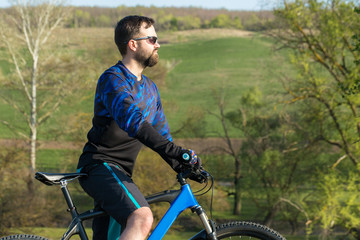 Cyclist in shorts and jersey on a modern carbon hardtail bike with an air suspension fork rides off-road on green hills near the forest	
