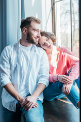cheerful woman with closed eyes sitting near window with handsome man at home