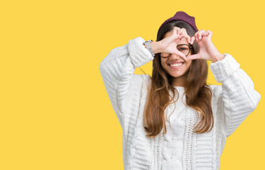 Young beautiful brunette hipster woman wearing glasses and winter hat over isolated background Doing heart shape with hand and fingers smiling looking through sign
