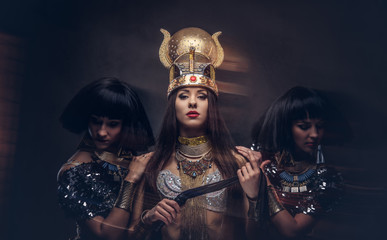 Fototapeta premium Portrait of haughty Egyptian queen in an ancient pharaoh costume with two concubines. Isolated on a dark background.