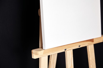 White blank cotton canvas stands on a wooden artistic easel on black curtain background. Horizontal rectangular mockup canvas wrapped on stretcher bar